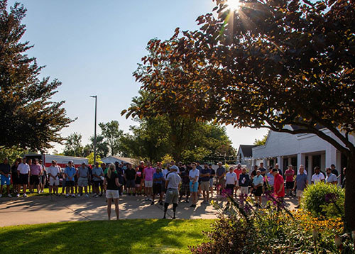 Large group of people standing outdoors, gathered around and listening to a person giving a speech.