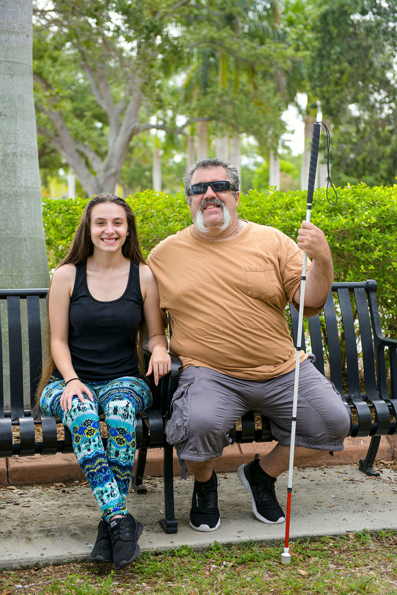 Aaron Cornelius, veterano herido, sosteniendo un bastón, sentado en un banco en un parque sonriendo con su hija Gabby.