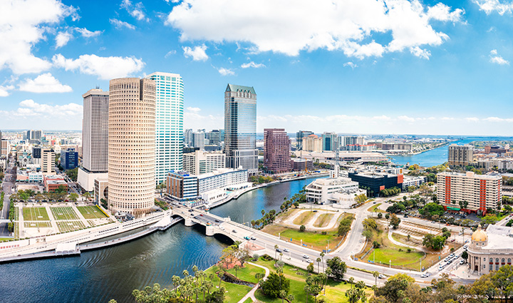 Aerial view of downtown Tampa with tall buildings, river and bridges, green spaces, and a mostly clear sky with scattered clouds. 