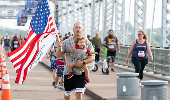 A man carries a flag and a baby in a chest carrier while running a Carry Forward 5K across a bridge.