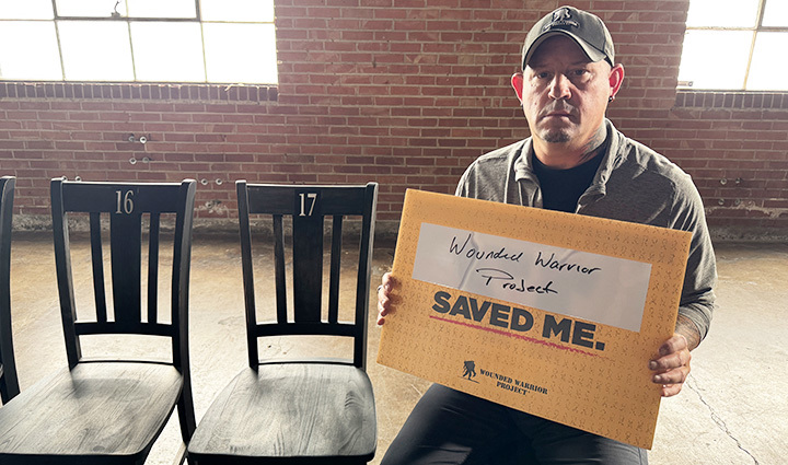 Warrior Phil Krabbe sits in a chair in front of a brick wall, holding a sign that says 