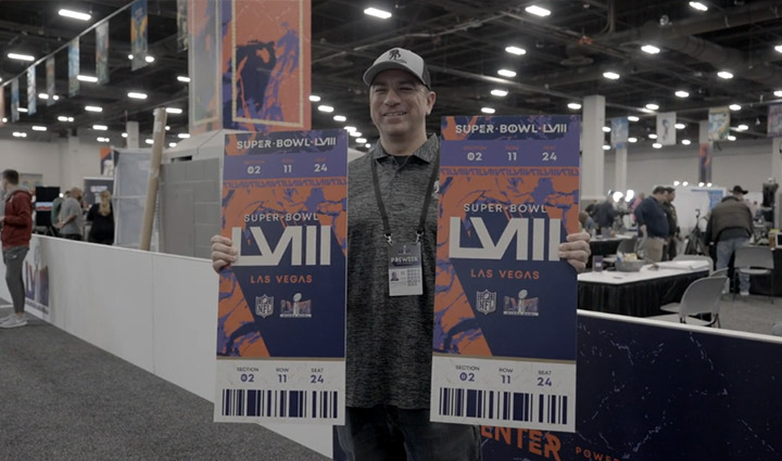 Wounded warrior Arnulfo Dauto stands in a convention hall holding two oversized Super Bowl tickets that were given to him by the NFL and USAA.