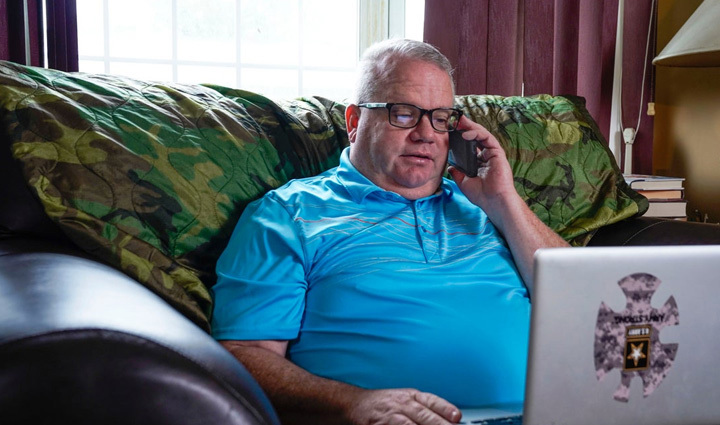 Warrior Chris Hoff talks on his phone while sitting on his couch in a blue polo working on his computer.