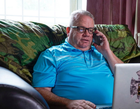 Warrior Chris Hoff talks on his phone while sitting on his couch in a blue polo working on his computer.