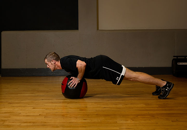 Wearing black athletic attire, wounded warrior John Rego maintains a low-plank pose while supporting himself on a fitness ball with his hands.