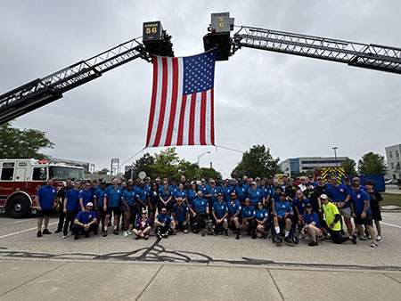 Participants gather for a large group photo under raised fire truck ladders displaying the American flag.