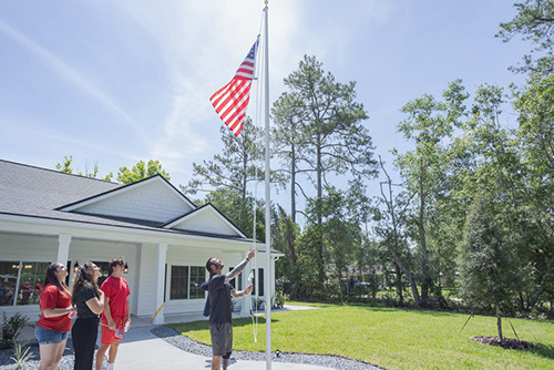 Sean Karpf stands outside his new home, looking up as he raises an American flag, while three people watch behind him.