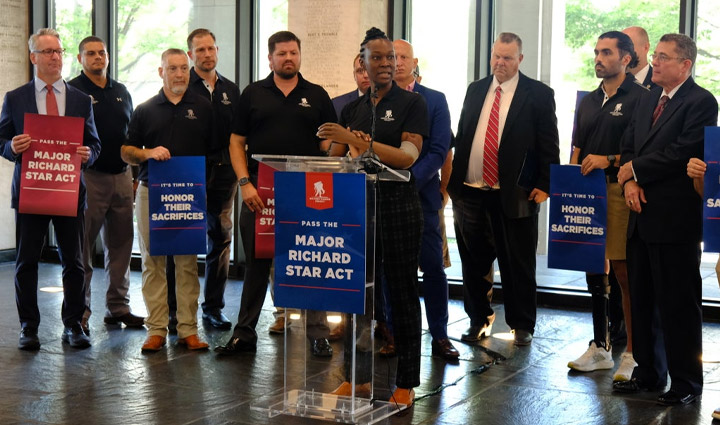 La veterana herida Danielle Green se encuentra en un podio con un micrófono y una pequeña multitud detrás de ella mientras habla a favor de la Ley del Mayor Richard Star.