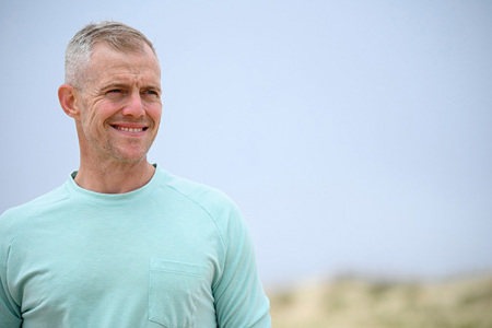 Army Ranger John Rego stands outdoors wearing a mint green long-sleeve shirt against a pale sky and sandy landscape.