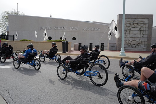A group of veterans ride recumbent bikes outside of the United States Naval Academy. They wear helmets and black Wounded Warrior Project shirts.
