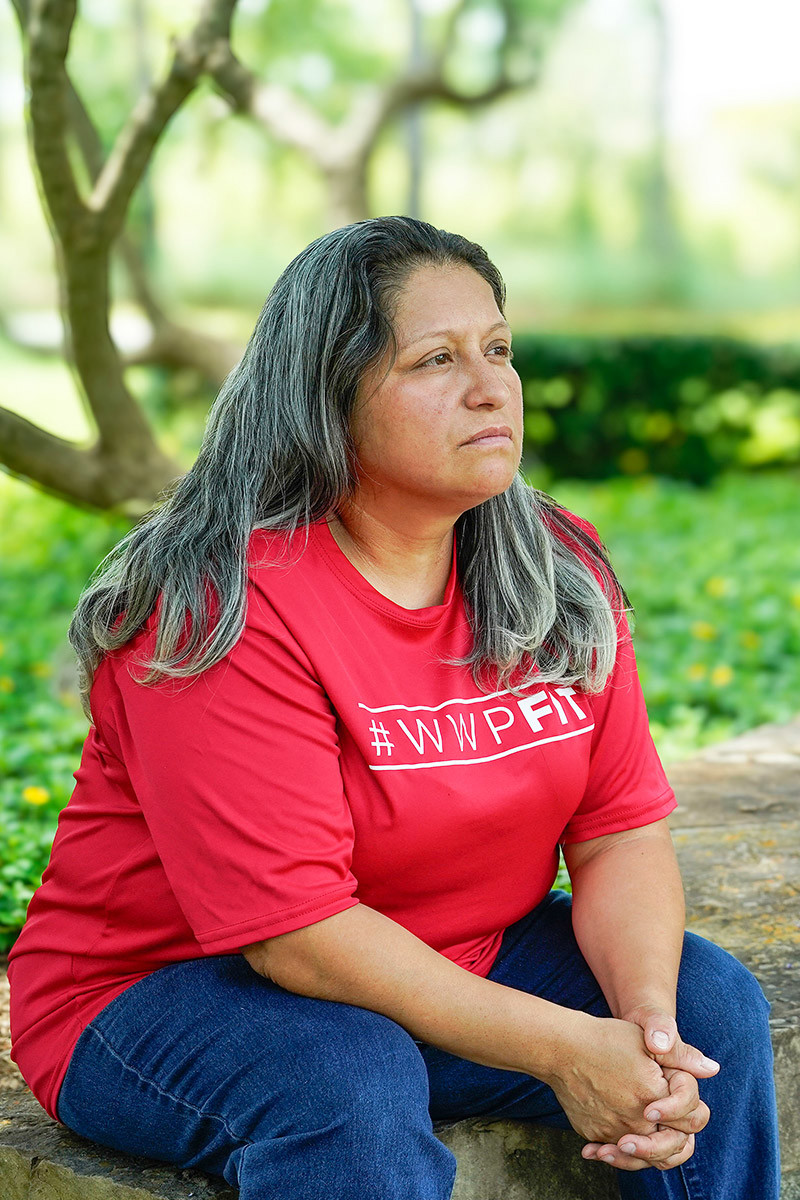 Wounded warrior Maria Edwards sits on a stone wall with trees in the background as she looks pensively off camera.