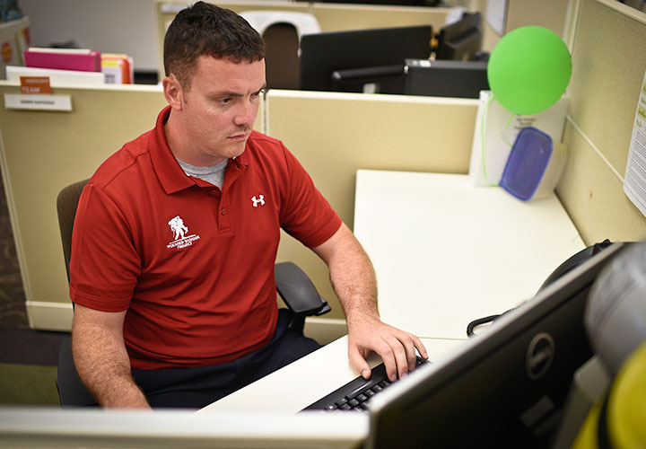 El veterano herido Jack Frawley usando una camiseta cuello polo roja de Wounded Warrior Project se sienta en un escritorio y trabaja diligentemente en una computadora.