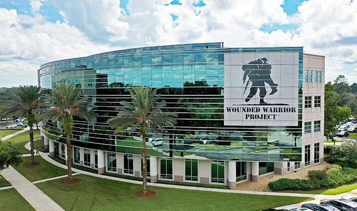Wounded Warrior Project HQ in Jacksonville, FL with a glass facade, logo, palm trees, and cars under a partly cloudy sky.