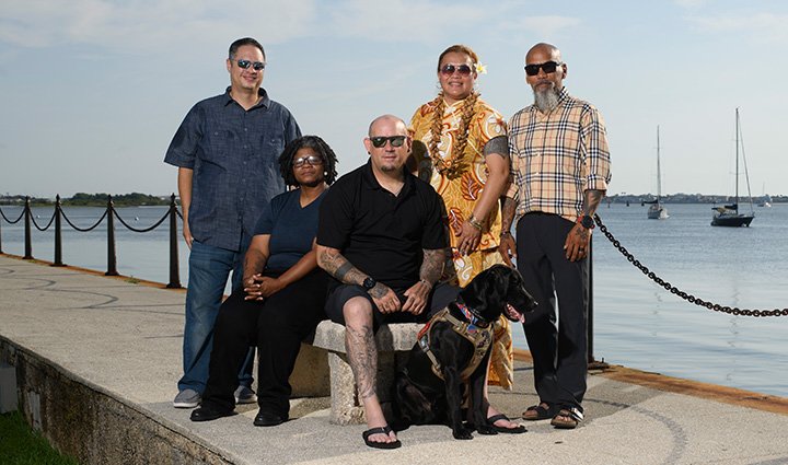 A group of five veterans, wounded warriors Jason Major, Pele Hunkin, Ray Andalio, Sam Hargrove, and Philip Krabbe and his black Labrador service dog pose for a picture at a waterfront with several boats in the background.