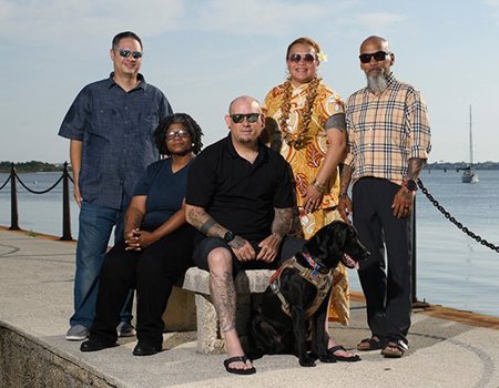 A group of five veterans, warriors Jason Major, Pele Hunkin, Ray Andalio, Sam Hargrove, and Philip Krabbe and his black Labrador service dog pose for a picture at a waterfront with several boats in the background.