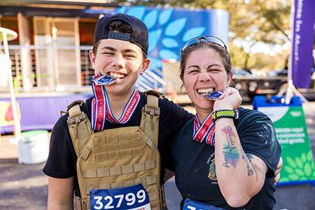 Two participants pose together at a Carry Forward event, smiling and playfully biting their medals.