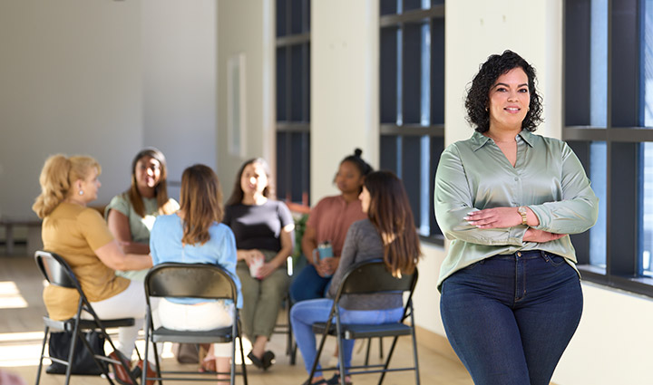 La veterana herida Yomari Cruz se encuentra de pie junto a un grupo de veteranas que hablan sentadas en círculo en el trabajo.