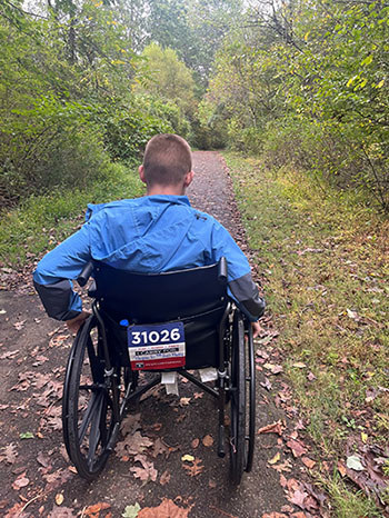 Carson Campos pushes himself forward in a wheelchair along an outdoor trail, with a Carry Forward 5K bib attached to the back.