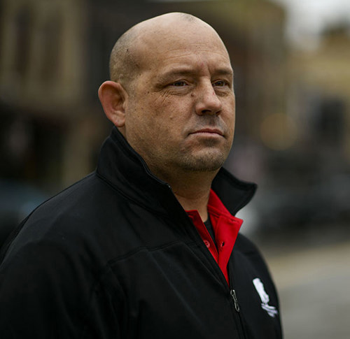 A serious-looking man in a black jacket with red collar stands outdoors. The background is blurred, suggesting an urban setting. The mood is contemplative.