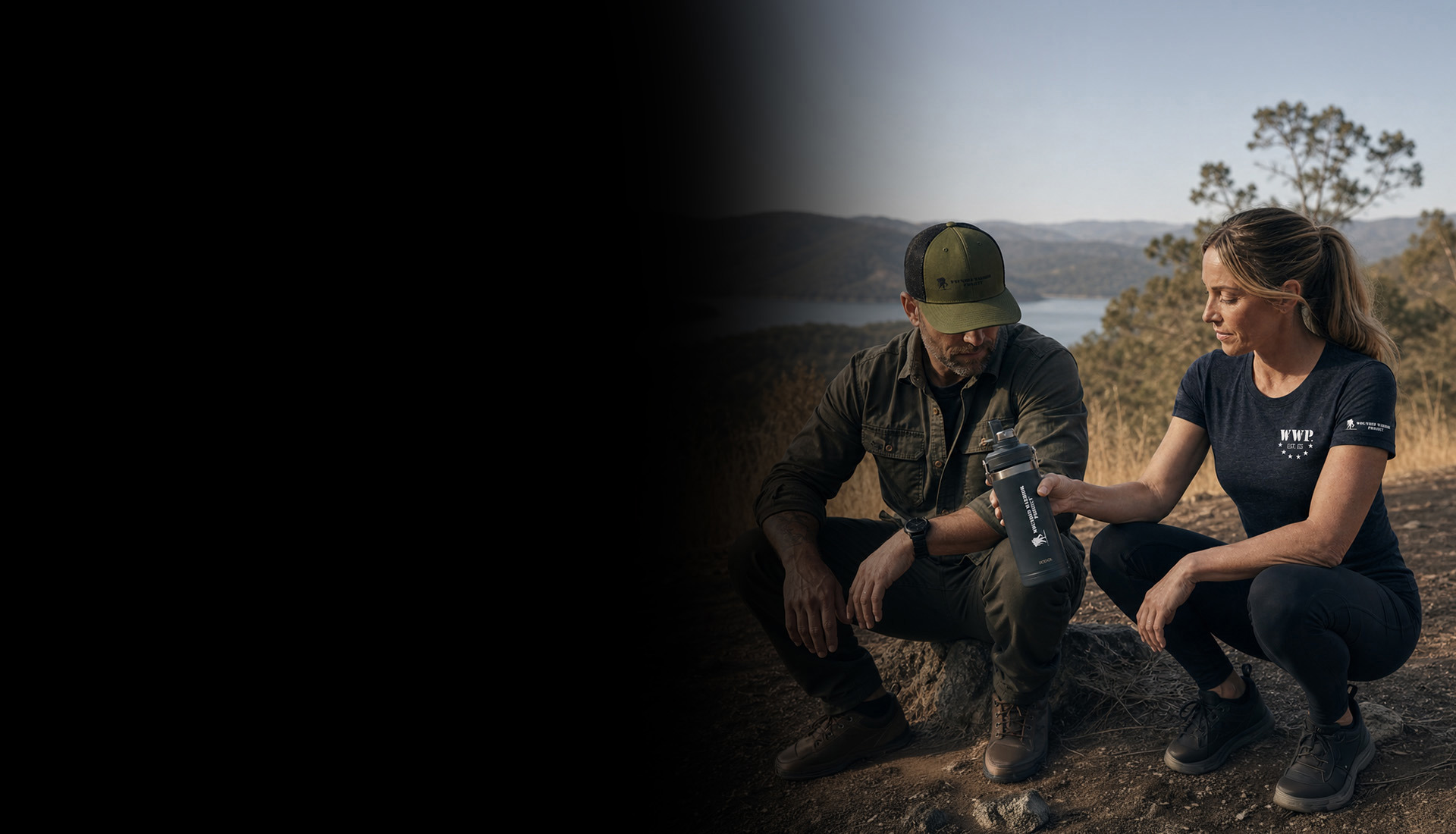 A man and woman wearing WWP-branded outdoor apparel crouch along a hiking trail overlooking a scenic lake and mountain landscape.