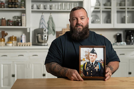 A military veteran sits at a kitchen table holding a framed portrait taken during military service. The framed photo shows the veteran in dress uniform as a younger service member. White kitchen cabinets and shelves are visible in the background.