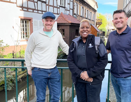 Warriors John Rego, Donna Pratt, and Mike Larson stand in front of a river in between two buildings while smiling.