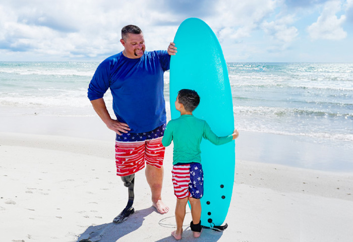 Wounded warrior Bryan Wagner with a prosthetic leg and his young son stand on a beach, holding a surf board together.