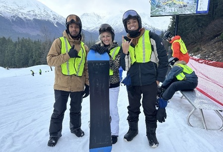 Three people in winter gear outside in snow, one holding a snowboard.