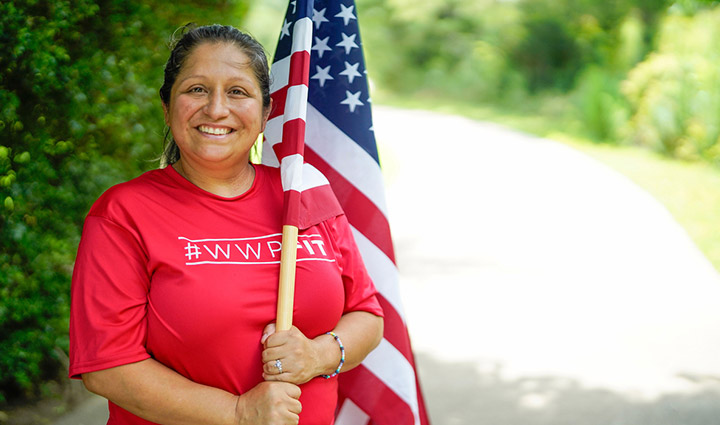 La veterana herida Maria Edwards usa una camiseta roja de WWP Fit mientras sonríe y sostiene una bandera estadounidense a lo largo de un camino arbolado.