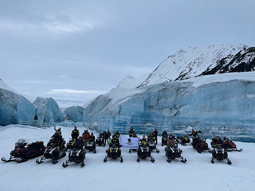 Group of people on snowmobiles, holding a WWP flag in front of a mountain.