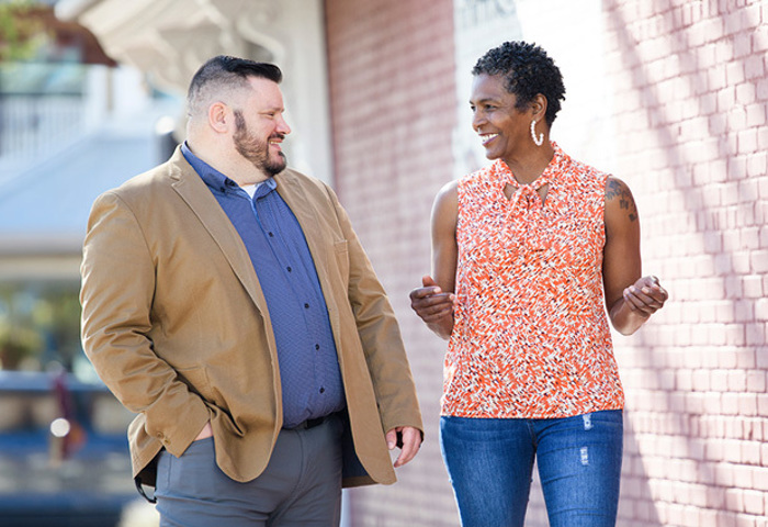 Warriors Jake Norotsky and Tonya Oxendine walk outside next to a brick wall while talking to each other and smiling.