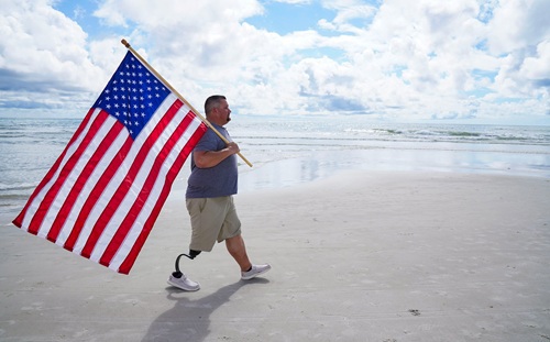 A man with a prosthetic leg walks on the beach carrying an American flag.
