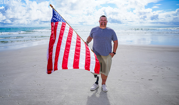 El veterano herido Bryan Wagner, tiene amputada una sola pierna, y está de pie en la playa con una bandera estadounidense.