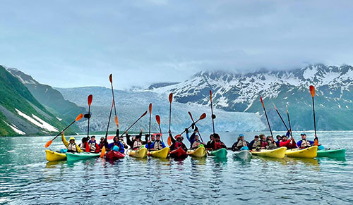 Group of kayakers in front of a mount, raising paddles in celebration.